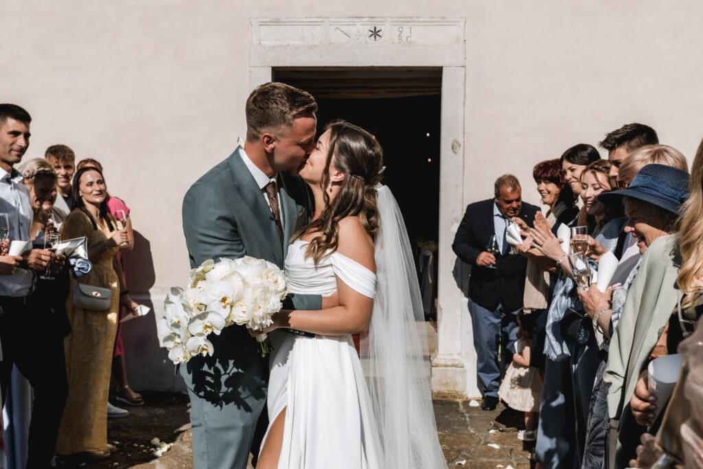 Mladoporočenca si pred cerkvijo delita poljub, medtem ko ju obkrožajo svatje. Nevesta v rokah drži eleganten poročni šopek iz belih orhidej in belih cvetov.